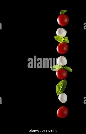 Vertikale Zusammensetzung von Mozzarella-Käse, frische Kirschtomate und Basilikumblätter isoliert auf schwarzem Hintergrund mit Reflexion, gesundes Lebensmittelkonzept, Stockfoto