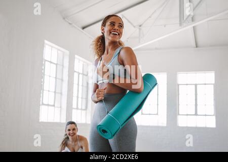 Lächelnde Fitness-Frau, die in einem Fitnessstudio steht und eine Yogamatte trägt. Porträt einer jungen Frau in einem Fitness-Trainingszentrum. Stockfoto