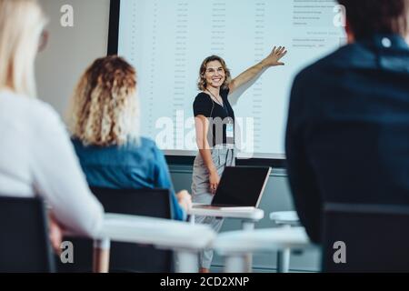 Geschäftsfrau hält eine Präsentation, während sie auf den Bildschirm zeigt, Kollegen ansieht und lächelt. Weibliche Führungskraft, die eine informative Präsentation gibt Stockfoto