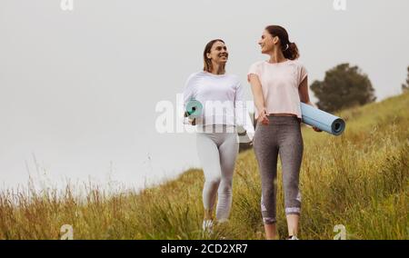 Lächelnde Freunde in Fitness tragen Walking in den Wiesen mit Yoga-Matten. Zwei Fitness-Frauen gehen für ein Training im Freien. Stockfoto