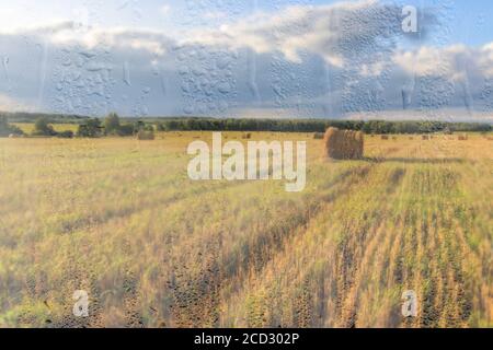 Ein landwirtschaftliches Feld mit Heuhaufen, beleuchtet von Abendsonne nach Regen, hinter nassem Glas im Herbst. Das Konzept der Änderung von schlechtem Wetter zu g Stockfoto