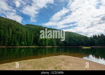 Landschaft rund um den See in den Bergen. Fichtenwald am Ufer. Spiegelung im Wasser. Sonniges Wetter mit Wolken am blauen Himmel Stockfoto