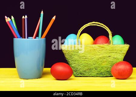 Osterkorb in grüner Farbe voller milticolor bemalter Eier, mehrere rote Eier auf Holztisch und blauer Becher mit Buntstiften isoliert auf schwarzem Hintergrund. Osterfeiertagskonzept Stockfoto