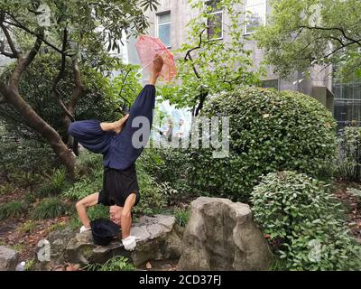 Herr Jiang, ein Mann in den Siebzigern, praktiziert Yoga auf besondere Weise, steht kopfüber mit dem Kopf und hält einen Schirm mit dem Fuß auf einem Ro Stockfoto