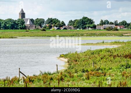 Zwolle, Niederlande, 21. Juli 2020: Flusskanal und Wildblumen in den Auen der IJssel mit der Kirche und den Häusern des Dorfes Stockfoto