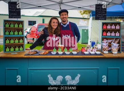 Junges französisches Paar in kastanienbraunen Schürzen vor ihrem Gewürzstand auf den Victor Harbor Farmers Markets in Südaustralien. Stockfoto