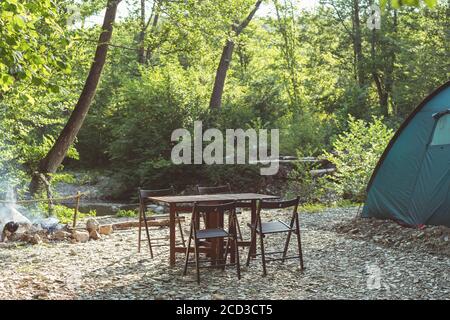 Camping am Strand des Bergflusses. Blaues Zelt, Lagertisch und Stühle im Wald. Sommerreisethema. Ruhestätte in der Natur. Stockfoto