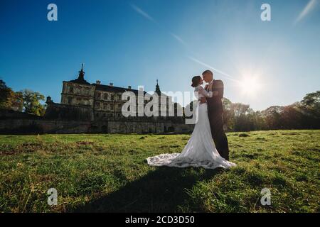 Schönes Hochzeitspaar, das im Sonnenlicht in der Nähe des alten Schlosses spazierenging. Stilvolle schöne Braut und Bräutigam in Armen vor dem Hintergrund eines alten Stockfoto