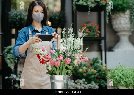 Blumenhändler bei der Arbeit. Frau in Schürze und Schutzmaske macht Foto auf Smartphone von Blumenstrauß Stockfoto