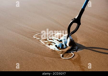 Nahaufnahme einer Person, die eine Reichweite ausdehnt, um eine zu sammeln Blau verwendet chirurgische Maske auf den feuchten Sand geworfen Die Küste oder zurück durch den Ozean zu Th gebracht Stockfoto