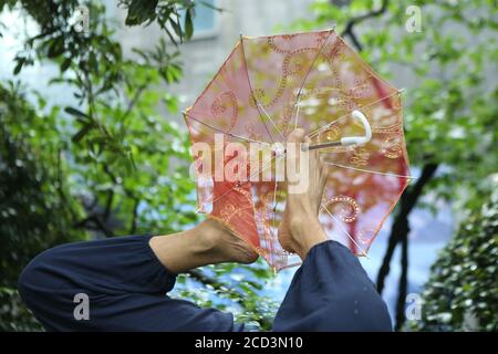 Herr Jiang, ein Mann in den Siebzigern, praktiziert Yoga auf besondere Weise, steht kopfüber mit dem Kopf und hält einen Schirm mit dem Fuß auf einem Ro Stockfoto