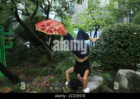 Herr Jiang, ein Mann in den Siebzigern, praktiziert Yoga auf besondere Weise, steht kopfüber mit dem Kopf und hält einen Schirm mit dem Fuß auf einem Ro Stockfoto