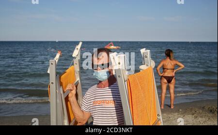 Rimini, Italien. Juli 2020. Ein Mann trägt eine Gesichtsmaske trägt Stühle am Strand während der Covid-19 Pandemie in Rimini, Italien. Quelle: Cezaro De Luca/dpa - ACHTUNG: Nur für redaktionelle Verwendung im Zusammenhang mit der aktuellen Berichterstattung und nur mit vollständiger Erwähnung der oben genannten Credit/dpa/Alamy Live News Stockfoto