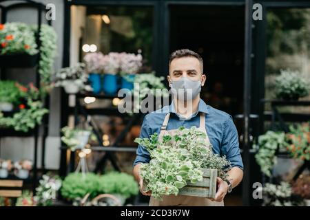 Kleinunternehmen und Beginn des Arbeitstages. Mann in Schutzmaske nimmt Kasten der Pflanzen draußen Stockfoto