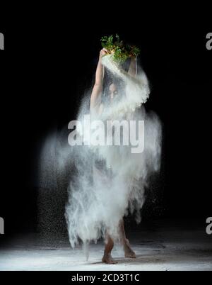 Schöne Frau mit kurzen schwarzen Haaren tanzt auf einem dunklen Hintergrund, hält ein Sieb mit Mehl. Sportlerin in schwarzem Bodysuit Stockfoto