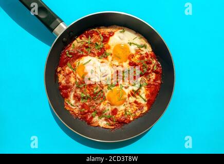 Shakshuka - pochierte Eier in Tomatensauce, Zwiebel, Pfeffer und Gewürze in eiserner Pfanne auf blauem hellen Hintergrund. Berühmte traditionelle arabische und israelische Frühstück Stockfoto