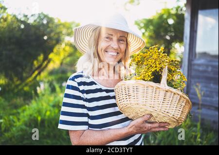 Glücklich lächelnd ältere Frau im Strohhut mit Spaß posieren im Sommergarten mit Blumen im Korb. Landwirtschaft, Gartenbau, Landwirtschaft, Rentner Stockfoto