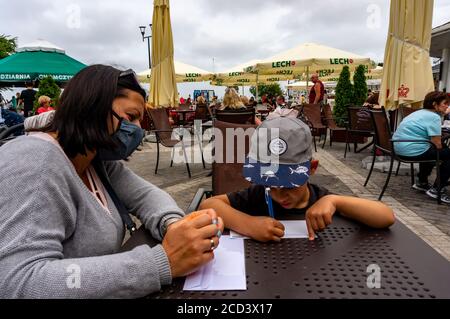 USTKA, POLEN - 03. Aug 2020: Frau mit Gesichtsmaske und Kind sitzt an einem Restauranttisch und schreibt eine Postkarte im Stadtzentrum. Stockfoto
