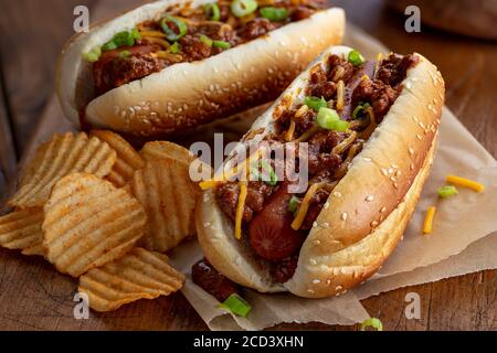 Chili Hot Dog mit Cheddar-Käse und grünen Zwiebeln auf Ein Sesambrötchen mit Kartoffelchips auf einem rustikalen Holzbrett Stockfoto