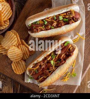 Chili Hot Dog mit Cheddar-Käse und grünen Zwiebeln auf Ein Sesambrötchen mit Kartoffelchips auf einem rustikalen Holzbrett Stockfoto