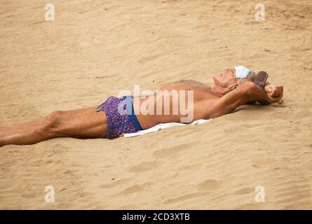 Las Palmas, Gran Canaria, Kanarische Inseln, Spanien. August 2020. Ein Sonnenanbeter trägt seine Gesichtsmaske auf seiner Stirn an ruhigen Tagen am Stadtstrand in Las Palmas auf Gran Canaria. Mit Covid 19 Fällen steigt auf Gran Canaria, hat die lokale Regierung verboten Joggen auf der Strandpromenade während des Tages sowie Sonnenbaden am Strand bei Flut zu verhindern, dass Gedränge. Im August wurden auf Gran Canaria mehr neue Coronavirus-Fälle registriert als in den Monaten März bis August zusammen. Mehr als 100 der neuen Fälle wurden auf eine Disco in der Hauptstadt Las Palmas zurückgeführt Stockfoto