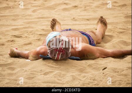 Las Palmas, Gran Canaria, Kanarische Inseln, Spanien. August 2020. Ein Sonnenanbeter trägt seine Gesichtsmaske auf seiner Stirn an ruhigen Tagen am Stadtstrand in Las Palmas auf Gran Canaria. Mit Covid 19 Fällen steigt auf Gran Canaria, hat die lokale Regierung verboten Joggen auf der Strandpromenade während des Tages sowie Sonnenbaden am Strand bei Flut zu verhindern, dass Gedränge. Im August wurden auf Gran Canaria mehr neue Coronavirus-Fälle registriert als in den Monaten März bis August zusammen. Mehr als 100 der neuen Fälle wurden auf eine Disco in der Hauptstadt Las Palmas zurückgeführt Stockfoto