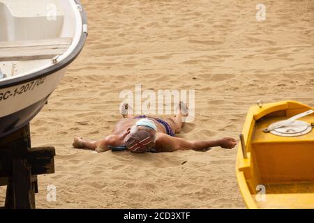 Las Palmas, Gran Canaria, Kanarische Inseln, Spanien. August 2020. Ein Sonnenanbeter trägt seine Gesichtsmaske auf seiner Stirn an ruhigen Tagen am Stadtstrand in Las Palmas auf Gran Canaria. Mit Covid 19 Fällen steigt auf Gran Canaria, hat die lokale Regierung verboten Joggen auf der Strandpromenade während des Tages sowie Sonnenbaden am Strand bei Flut zu verhindern, dass Gedränge. Im August wurden auf Gran Canaria mehr neue Coronavirus-Fälle registriert als in den Monaten März bis August zusammen. Mehr als 100 der neuen Fälle wurden auf eine Disco in der Hauptstadt Las Palmas zurückgeführt Stockfoto