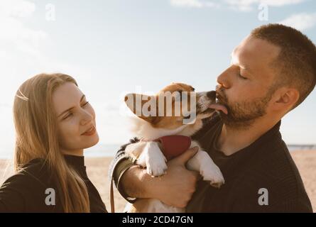 Junges glückliches Paar mit Hund macht Selfie-Foto am Strand. Schöne Familie und Corgi Welpen lecken Mann Wange. Familie mit Haustier im Freien, gemeinsam zu Fuß, Mann und Frau entspannen im Sommer am Meer Stockfoto