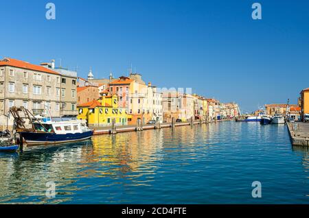 Schmaler Wasserkanal mit Booten und Fischerschiffen, die in der Nähe von Böschungen und bunten Gebäuden im historischen Zentrum der Stadt Chioggia festgemacht sind, blauer Himmel im Sommer, Region Venetien, Norditalien Stockfoto