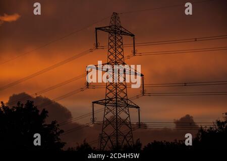 Strommasten füttern die lokale Gegend in West Wales an einem Abend, die die Sonnenuntergänge ein tiefes Orange zwischen den Wolken. Stockfoto