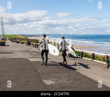 Zwei junge Männer in nassen Anzügen tragen ihre Surfbretter Entlang der Promenade in Saltburn by the Sea, England, Großbritannien Stockfoto