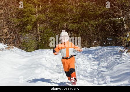 Kleiner Junge in einem orangefarbenen Jumpsuit, der auf schneebedeckter Straße in einem Nadelwald läuft. Sonniger Wintertag Stockfoto