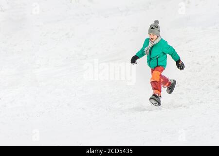 Boy in der grünen Jacke und roten Hose läuft durch den Schnee. Wintertag. Stockfoto