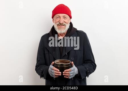 Reifer lächelnder Po in schwarzem Mantel und roter Mütze auf dem Kopf mit Eisenbügelbüchsenhalter für die Geldbeschaffung. Isoliert auf weißem Hintergrund Stockfoto