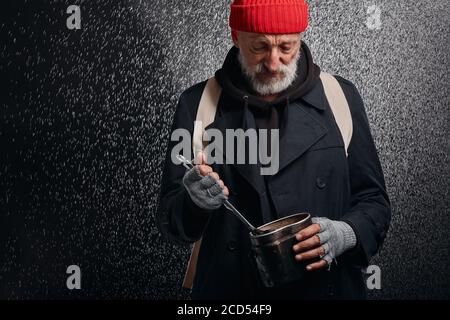 Älterer graubärtiger Mann mit eiserner Pfanne und Suppenkelle. Versuchen Sie, etwas Essen auf dem Boden der Pfanne zu finden. Isoliert auf schwarzem Hintergrund Stockfoto