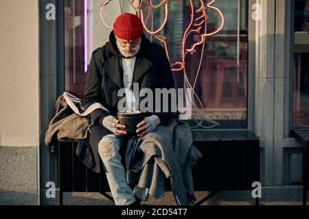 Reifer kaukasischer vagrant Mann bitten um einige Geldspenden von Bürgern, hoffentlich Blick auf rostigen leeren Glas. Modernes Café hinter ihm. Armut, Hunger, h Stockfoto