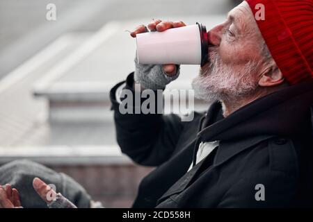 Älterer Bettler mit grauem Bart, der Tee trinkt. Armer Mann mit alten schmutzigen Kleidern ist hungrig. Seitenansicht Stockfoto