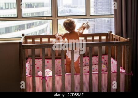 Stay Home Konzept. Baby Kleinkind mit Spielzeug Katze steht in der Krippe und Blick aus dem Fenster auf die Wolkenkratzer Stadtbild Stockfoto