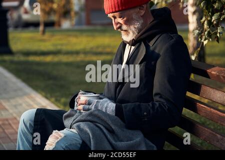 Reifer Mann ohne Unterschlupf sitzt in Straßenkleidung auf der Bank, ohne Essen und Geld. Der herabhängende Mann senkte seinen Kopf nach unten. Seitenansicht auf Obdachlose Perso Stockfoto