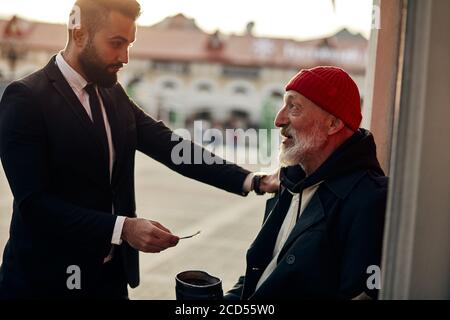 Geschäftsmann in Smoking helfen, vagrant Mann auf der Straße mit rostigen Geldkrug sitzen. Der reiche Mann legte eine Hand auf die Schulter der Obdachlosen und gab einem zu tun Stockfoto