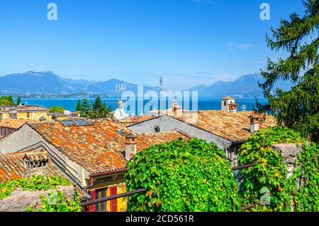 Luftaufnahme von Desenzano del Garda Stadt mit roten Ziegeldachgebäuden, grünen Pflanzen, Gardasee Wasseroberfläche, Monte Baldo Bergkette, blauer Himmel Hintergrund, Lombardei, Norditalien Stockfoto