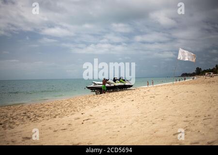 Man lädt auf einem Trolley Jet Skis auf einem Sandstrand auf einem Hintergrund des Meeres mit Wellen. Eine weiße Flagge winkt in der Nähe mit einem Platz für einen Speer des Raumes Stockfoto