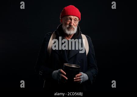 Alter obdachloser Mann in rotem Hut und grauen Strickhandschuhen, hält Ironie Konserve Bank jar für Geld Almosen in Studio schwarz Hintergrund Stockfoto