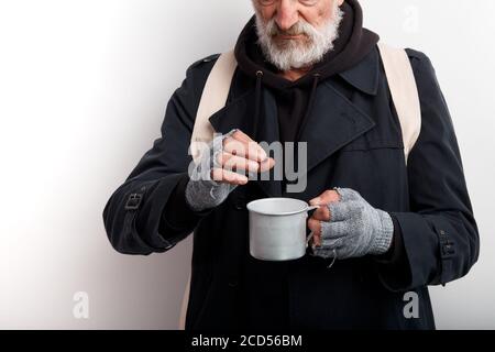 Obdachloser, reifer bärtiger Mann in schwarzem Mantel und weißem Rucksack, der Tasse für die Geldbeschaffung hält. Isoliert auf weißem Hintergrund Stockfoto