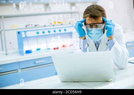Hübscher junger Forscher in weißem Labormantel und Schutzmaske Arbeiten mit einem Laptop im Labor Stockfoto