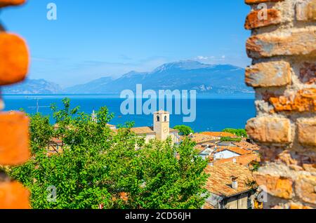 Blick auf das historische Zentrum Desenzano del Garda Altstadt mit Glockenturm, See und Bergkette durch Fenster aus Backsteinmauer der mittelalterlichen Burg, blauer Himmel Hintergrund, Lombardei, Norditalien Stockfoto