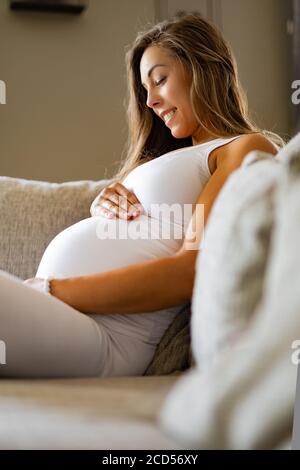 Lächelnd schwangere Frau sitzt im Sofa mit Blick auf ihren Bauch Stockfoto
