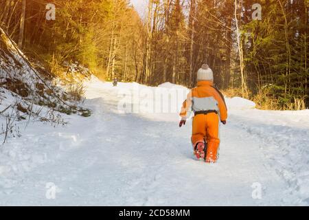 Kleiner Junge in einem orangefarbenen Jumpsuit, der auf schneebedeckter Straße in einem Nadelwald läuft. Mutter ist in der Ferne. Sonniger Wintertag. Rückansicht. Stockfoto