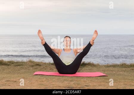 Frau in Sportbekleidung Pilates im Freien auf einer rosa Matte mit dem Meer im Hintergrund. Stockfoto
