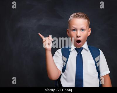 Überrascht kaukasischen Schuljunge zeigt Kopie Raum. Hübscher junger Mann im Hemd, der auf die Kamera schaut und wegzeigt, während er gegen die schwarze Schule steht Stockfoto